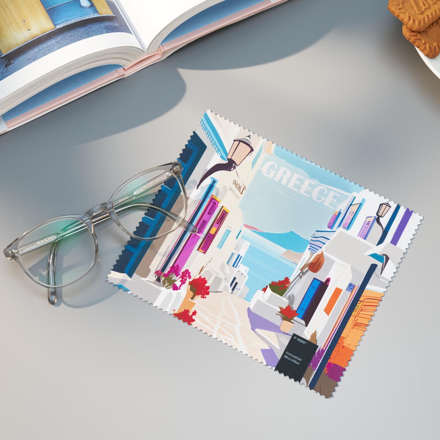 Ein quadratisches P-9000-Mikrofaser-Reinigungstuch mit einer farbenfrohen Griechenland-Reiseillustration neben einer durchsichtigen Brille.