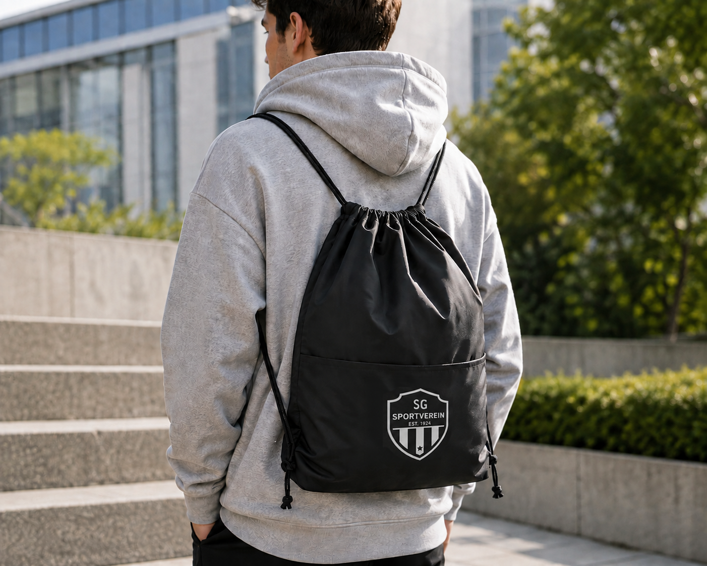 Eine Person trägt einen grauen Kapuzenpullover und eine schwarze Kordelzugtasche mit weißem SG-Sportverein-Logo im Freien an einem sonnigen Tag.
